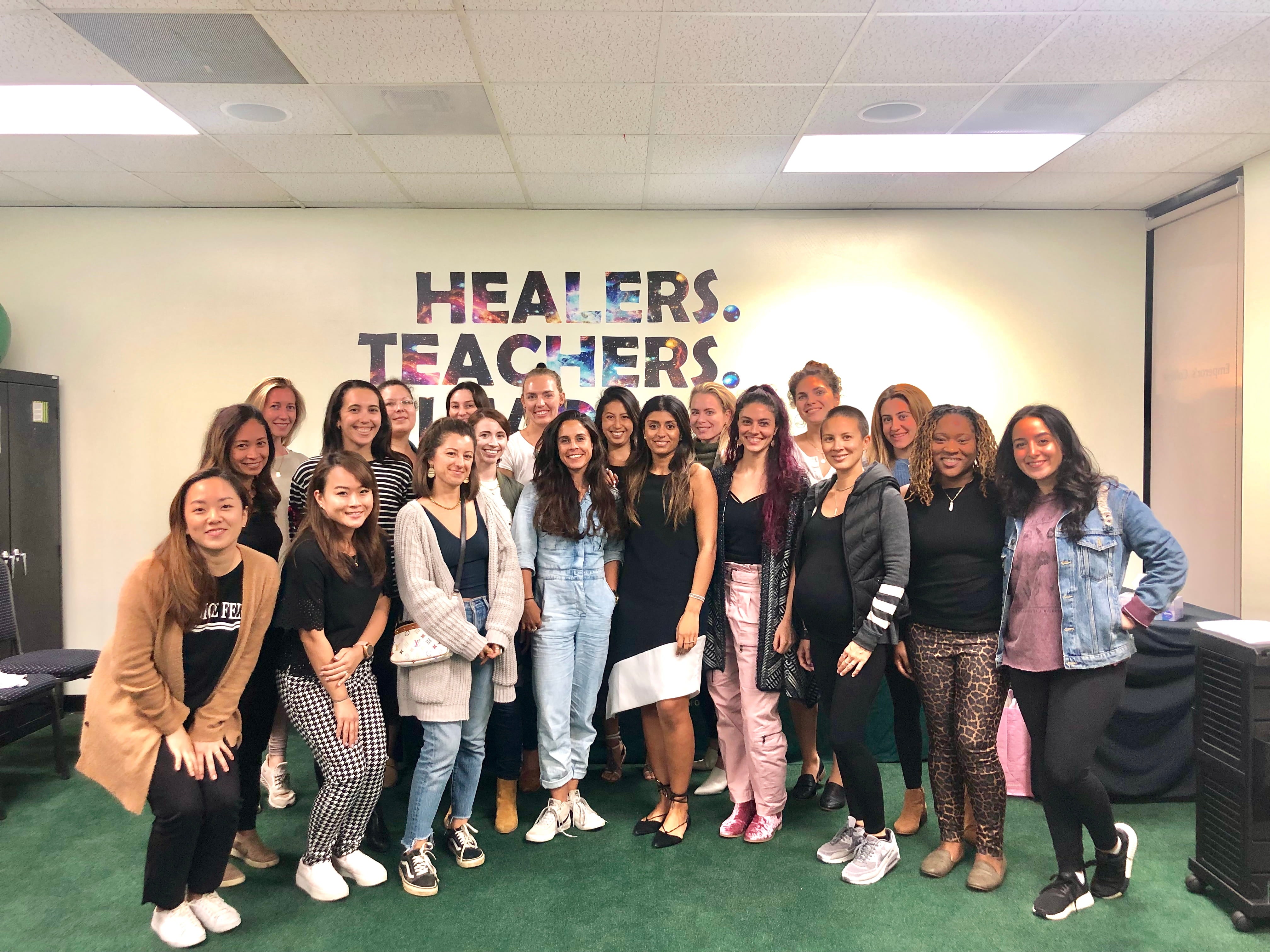 A group of 20 people, mostly women, stand together and smile in a classroom. Behind them, a colorful sign reads HEALERS. TEACHERS. The cheerful group shares a passion for Traditional Chinese Medicine skincare and learning new wellness practices.
