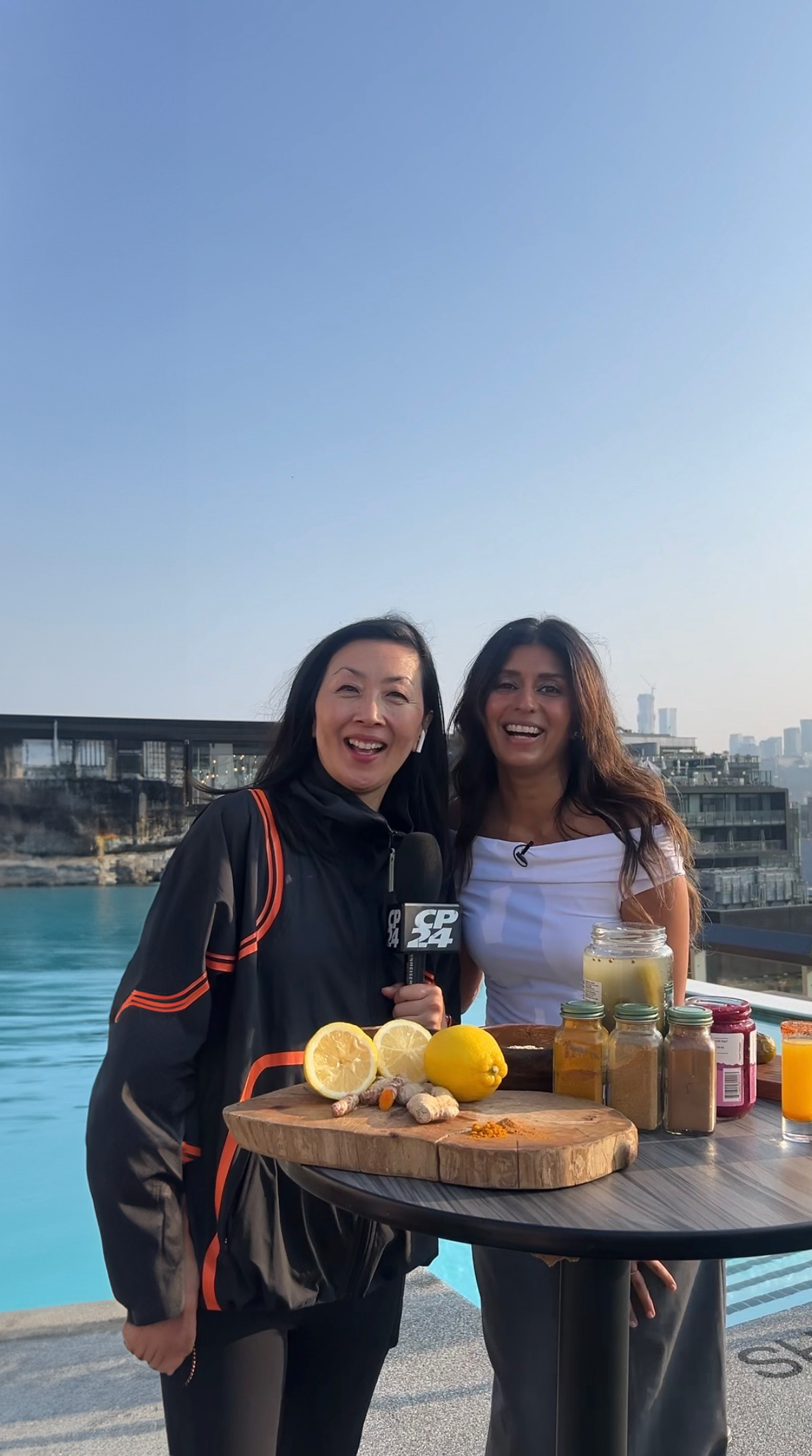Two women standing outdoors by a pool with a city skyline in the background.